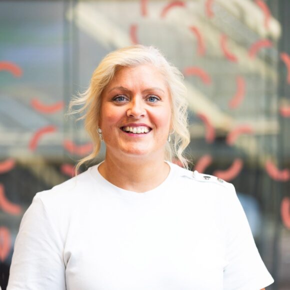 Gemma Dunn's headshot sees her wearing a white t-shirt standing against a glass background with coral squiggles