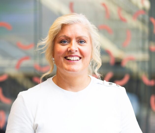 Gemma Dunn's headshot sees her wearing a white t-shirt standing against a glass background with coral squiggles