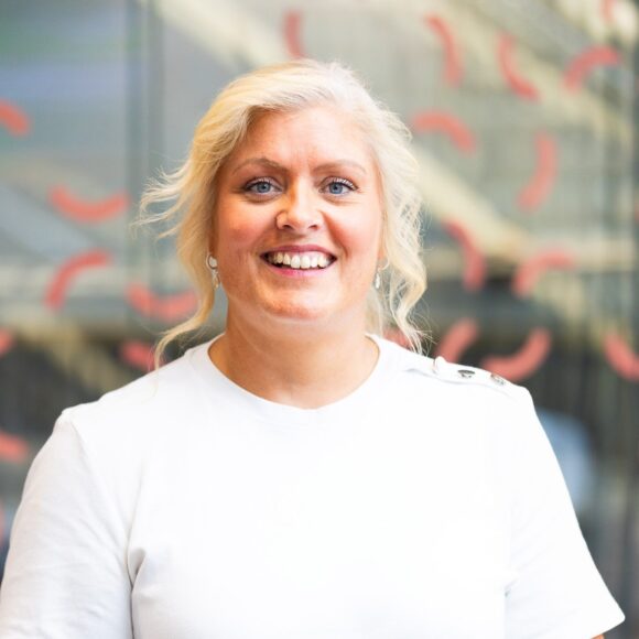 Gemma Dunn's headshot sees her wearing a white t-shirt standing against a glass background with coral squiggles