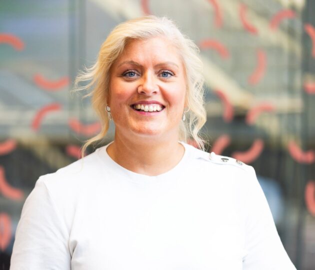 Gemma Dunn's headshot sees her wearing a white t-shirt standing against a glass background with coral squiggles